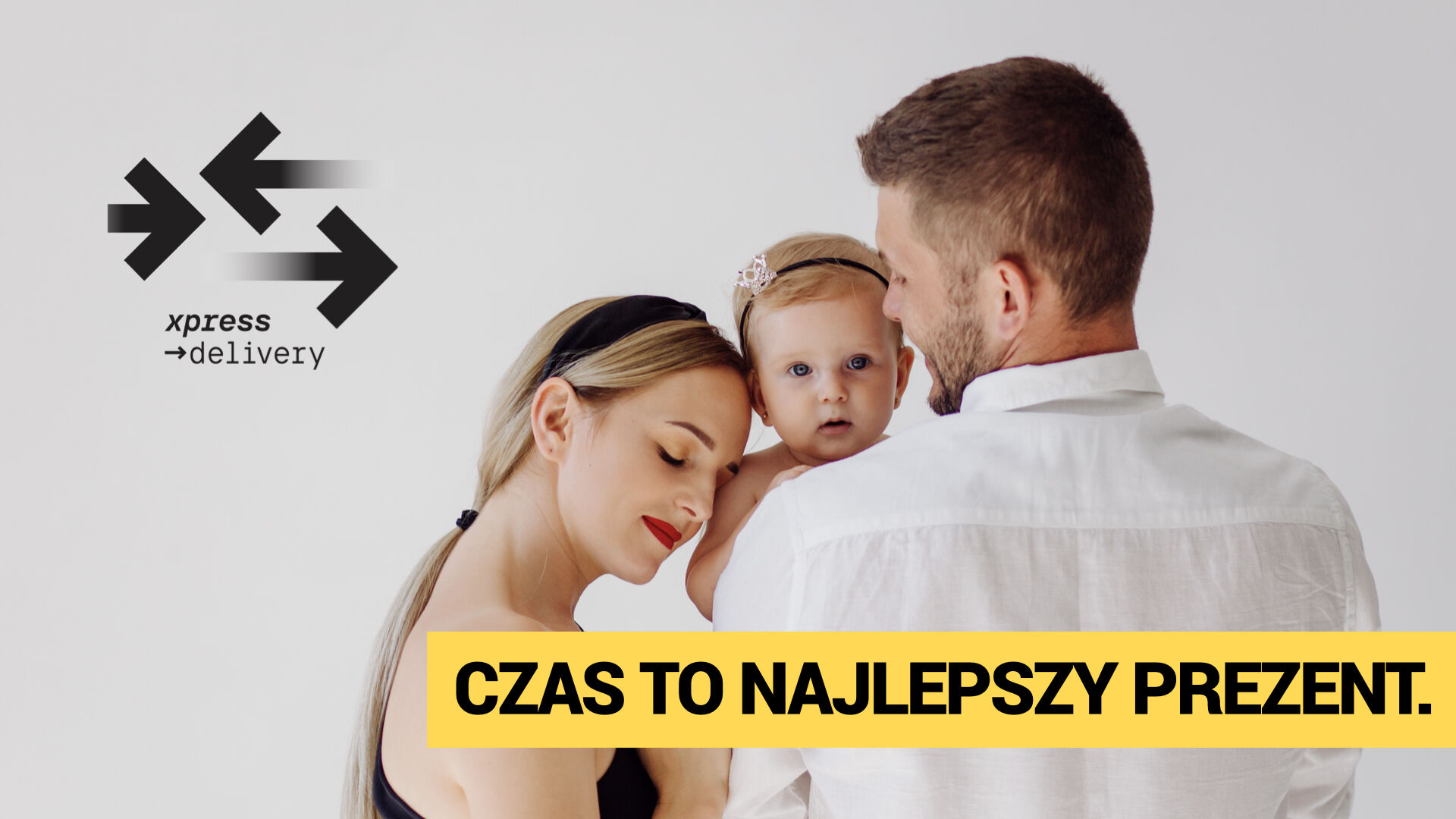 Młoda kobieta i mężczyzna trzymający niemowlę w objęciach, stoją razem na jasnym tle. Kobieta z zamkniętymi oczami i uśmiechem opiera głowę na ramieniu partnera. Dziecko patrzy prosto w obiektyw. W lewym górnym rogu widoczne jest logo xpress→delivery, a na dole, na żółtym pasku – wyrazisty napis: „CZAS TO NAJLEPSZY PREZENT.”
